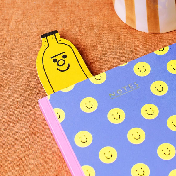 A yellow banana-shaped bookmark with a smiling face is tucked under the edge of a blue notebook covered in yellow smiley faces, set on an orange textured fabric surface.