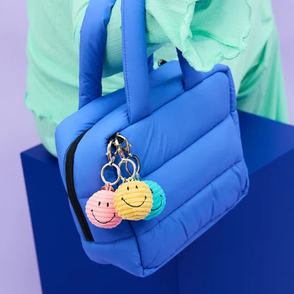 A bright blue puffer-style handbag with padded handles and a black zip. The bag has three colourful smiley-face keyring charms attached on the front, and the scene is set against a purple and blue studio background.