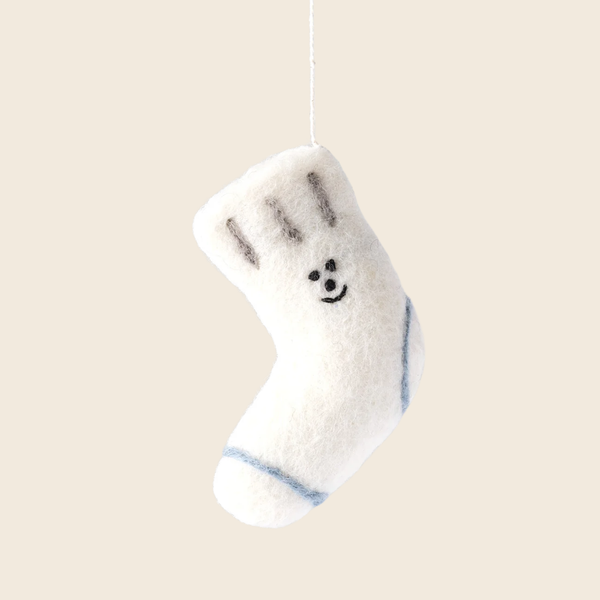 A small white felt Christmas stocking ornament hanging from a thin white string against a solid light beige background. The stocking features a simple black embroidered face and light blue stripes near the toe.