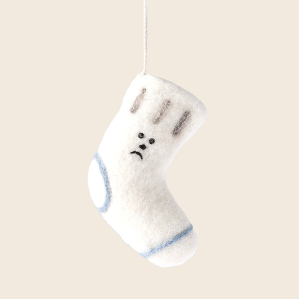A white felt Christmas stocking ornament with a sad face, hanging from a thin white string against a solid beige background.