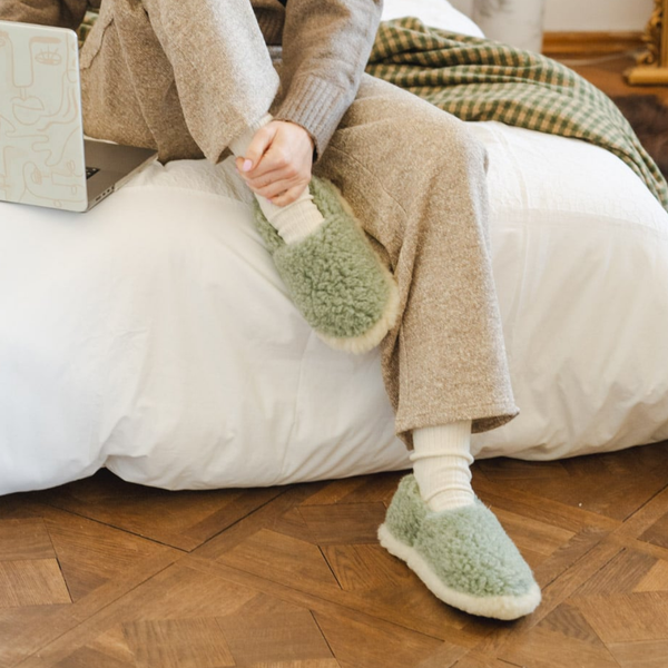 A person sitting on a white bed wearing cozy light green fuzzy slipper slippers with white socks in a warm, home-like setting with a wooden parquet floor.
