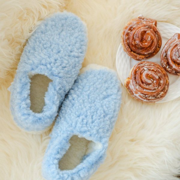 A pair of fluffy light blue slip-on slippers with cream lining, arranged on a cream faux-fur surface next to a white plate with three cinnamon rolls.