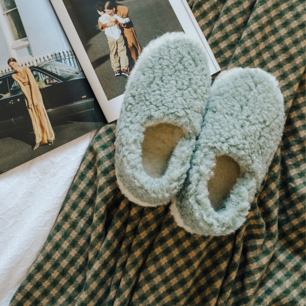 A pair of fluffy pale green slip-on slippers placed on a green and beige plaid blanket, with an open magazine visible in the background.